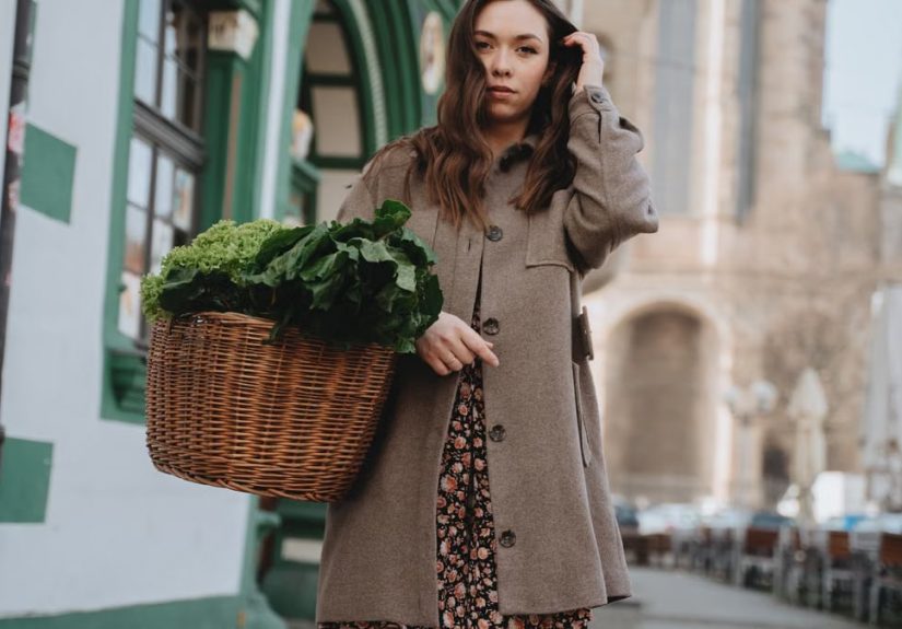 A woman in a brown coat carries a wicker basket of green vegetables while strolling through Erfurt, Germany.